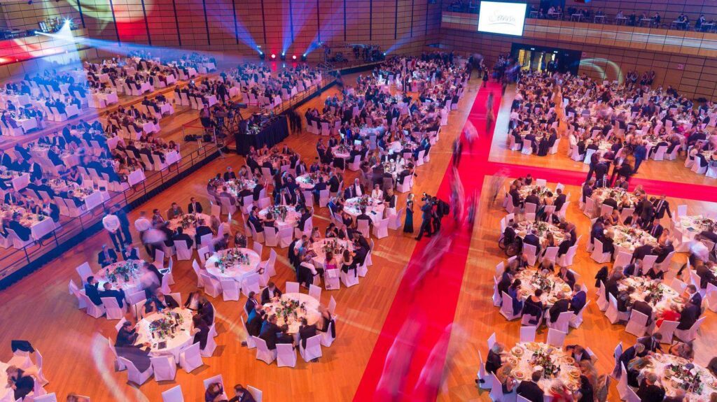 Formal gala dinner with round tables and red carpet in large hall.