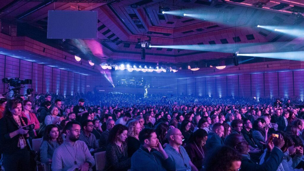 Large audience seated in a conference hall with stage lighting.
