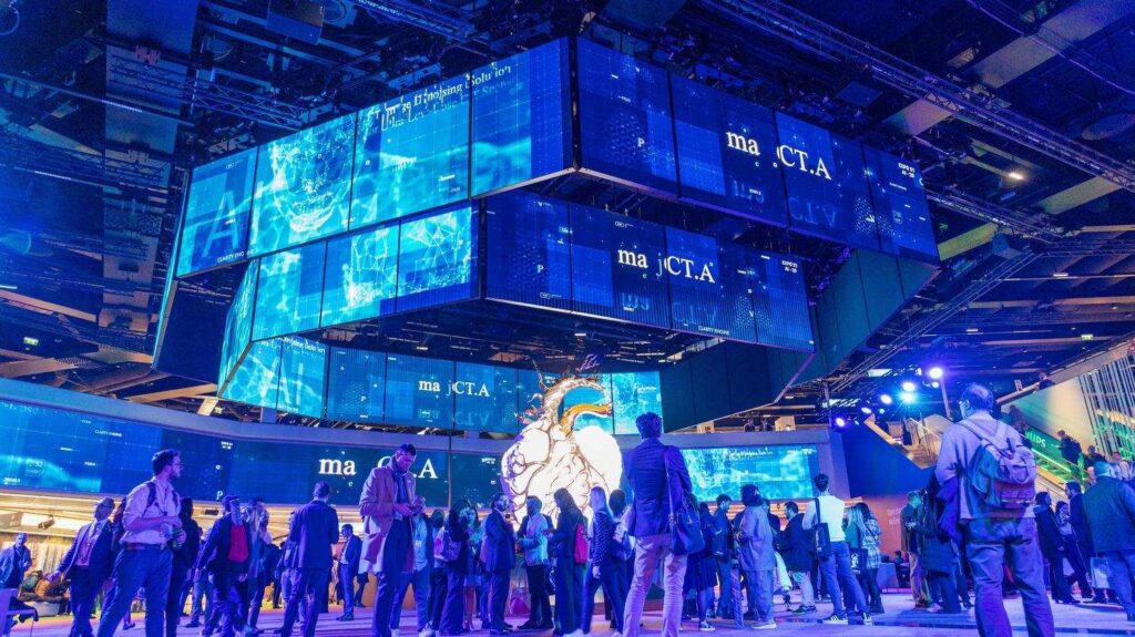 Visitors in exhibition hall beneath large suspended digital screens.