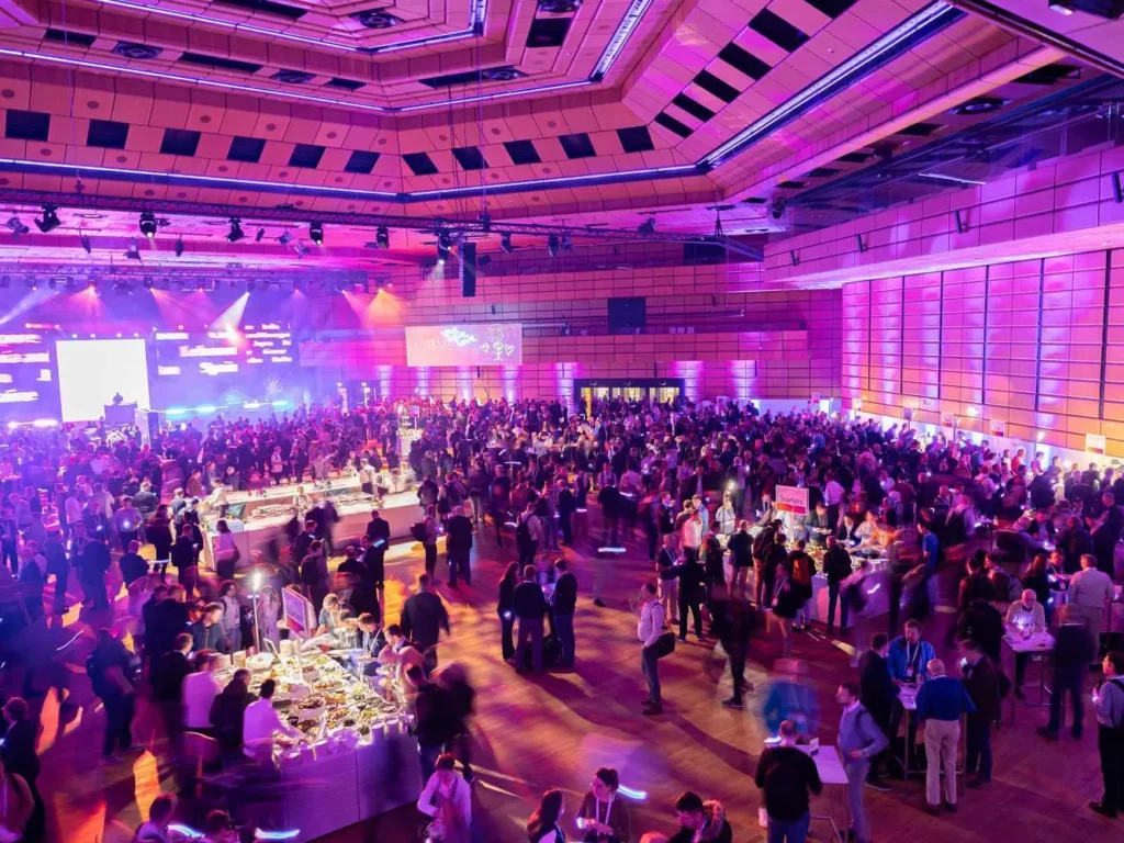 Large event hall with many guests, buffet areas and purple lighting.